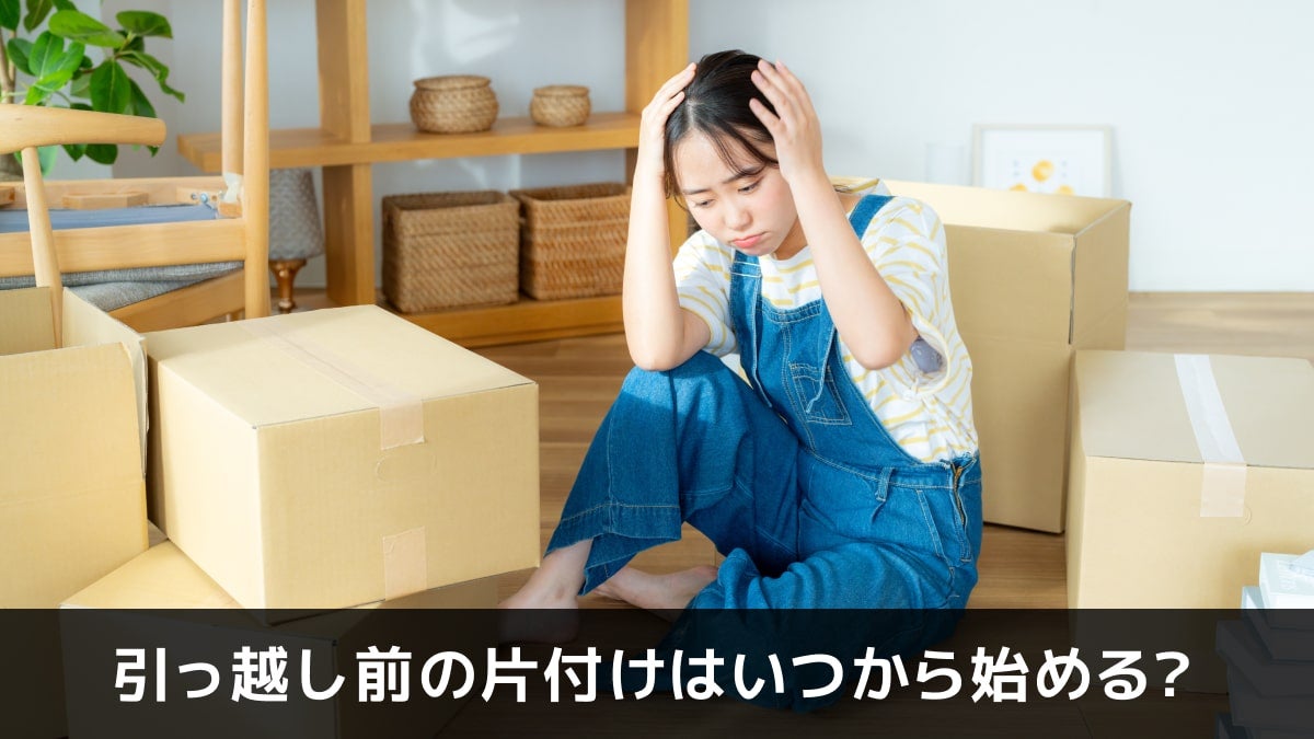 写真：引っ越し前の片付けはいつから始める？。ダンボールに囲まれた部屋で頭をかかえる女性の写真。