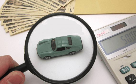 写真：車・バイクの輸送でかかる相場料金。お金と電卓と虫眼鏡で覗かれたミニチュアの車。